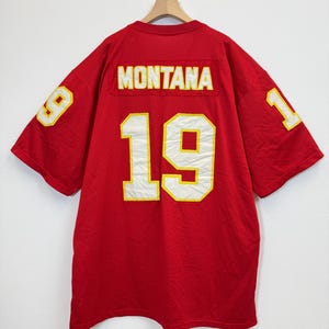 Puede incluir: Camiseta de malla roja con el nombre "MONTANA" en la parte superior de la espalda y el número "19" en blanco con ribete amarillo en la espalda y las mangas. La camiseta está colgada de una percha de madera.