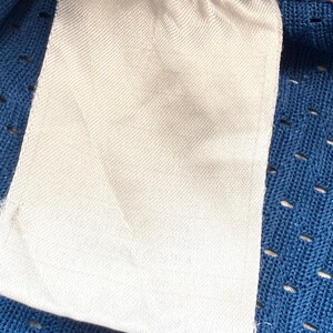 K&ouml;nnte beinhalten: Nahaufnahme eines wei&szlig;en Stoffetiketts mit leerer Oberfl&auml;che, befestigt an einem blauen Mesh-Gewebe. Das Etikett ist rechteckig mit abgerundeten Ecken und einer strukturierten Oberfl&auml;che. Der blaue Stoff hat kleine, gleichm&auml;&szlig;ig verteilte L&ouml;cher.