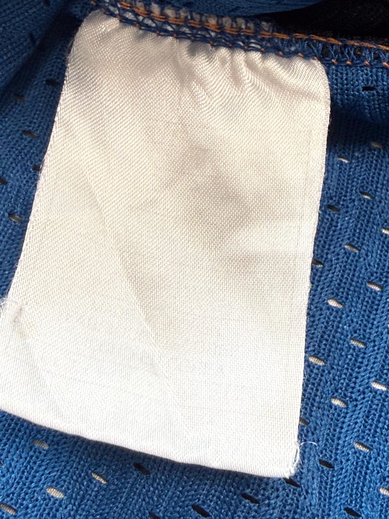 K&ouml;nnte beinhalten: Nahaufnahme eines wei&szlig;en Stoffetiketts mit leerer Oberfl&auml;che, befestigt an einem blauen Mesh-Gewebe. Das Etikett ist rechteckig mit abgerundeten Ecken und einer strukturierten Oberfl&auml;che. Der blaue Stoff hat kleine, gleichm&auml;&szlig;ig verteilte L&ouml;cher.