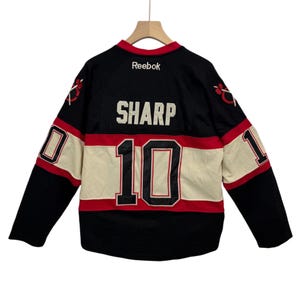 May include: Black hockey jersey with the name "SHARP" and the number "10" in white and red. The jersey has a cream-colored stripe across the chest and sleeves, with red and black accents. The Reebok logo is visible at the neckline.