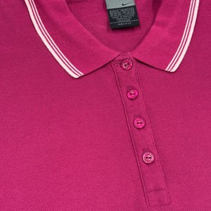 May include: A magenta polo shirt with a white and light pink striped collar. The shirt has a three-button placket and a small Nike tag near the collar. The fabric appears to be a breathable, athletic material.