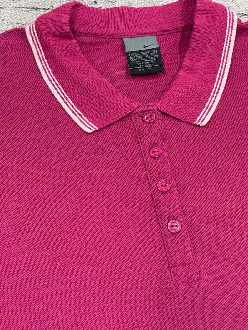 May include: A magenta polo shirt with a white and light pink striped collar. The shirt has a three-button placket and a small Nike tag near the collar. The fabric appears to be a breathable, athletic material.