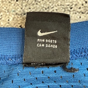 K&ouml;nnte beinhalten: Nahaufnahme eines schwarzen Nike-Etiketts mit wei&szlig;em Swoosh-Logo, RN# 94878 und CA# 34428. Das Etikett ist an einem blauen, perforierten Stoff befestigt. Der Hintergrund ist strukturiert und neutral gef&auml;rbt.