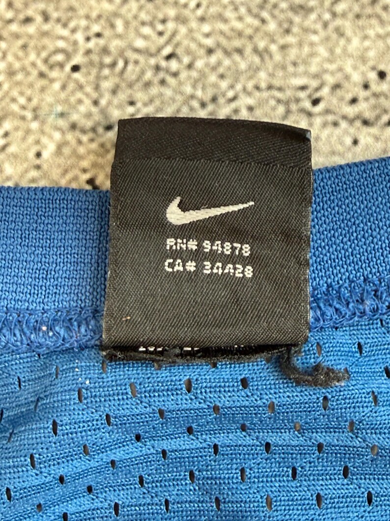 K&ouml;nnte beinhalten: Nahaufnahme eines schwarzen Nike-Etiketts mit wei&szlig;em Swoosh-Logo, RN# 94878 und CA# 34428. Das Etikett ist an einem blauen, perforierten Stoff befestigt. Der Hintergrund ist strukturiert und neutral gef&auml;rbt.