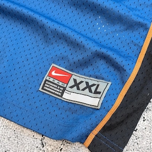 K&ouml;nnte beinhalten: Nahaufnahme einer blauen Mesh-Sporthose. Die Hose hat einen schwarzen und orangefarbenen Besatz an der Seite. Ein Nike-Etikett ist sichtbar, mit dem Text "TEAM ENGINEERED TO THE EXACT SPECIFICATIONS OF CHAMPIONSHIP ATHLETES" und der Gr&ouml;&szlig;e "XXL".