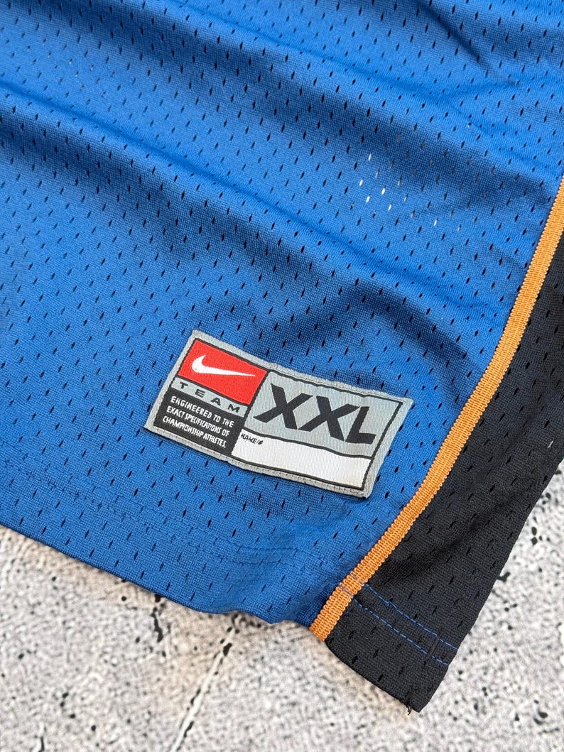 K&ouml;nnte beinhalten: Nahaufnahme einer blauen Mesh-Sporthose. Die Hose hat einen schwarzen und orangefarbenen Besatz an der Seite. Ein Nike-Etikett ist sichtbar, mit dem Text "TEAM ENGINEERED TO THE EXACT SPECIFICATIONS OF CHAMPIONSHIP ATHLETES" und der Gr&ouml;&szlig;e "XXL".