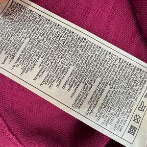 May include: A close-up of a care label on a magenta fabric. The white label has text in multiple languages, including washing instructions and symbols. The fabric has a textured appearance.