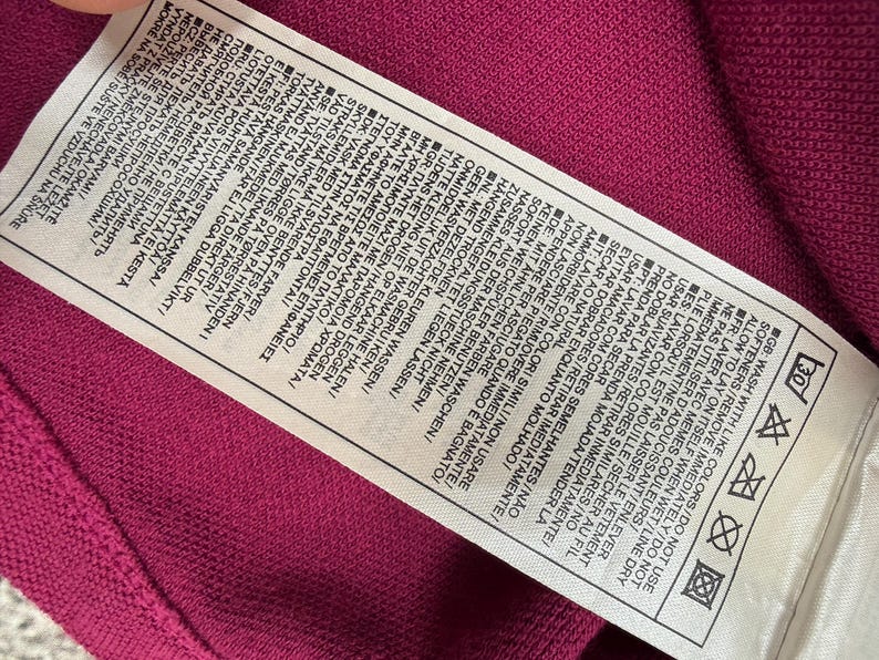 May include: A close-up of a care label on a magenta fabric. The white label has text in multiple languages, including washing instructions and symbols. The fabric has a textured appearance.