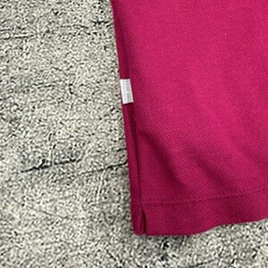 May include: Close-up of a magenta polo shirt. The fabric has a textured appearance. A small white tag is visible on the side seam. The shirt is made of a breathable material.