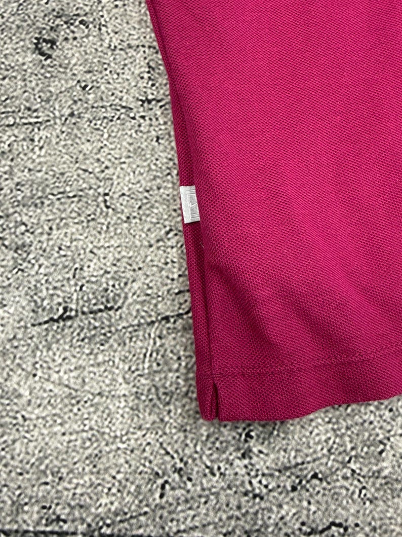 May include: Close-up of a magenta polo shirt. The fabric has a textured appearance. A small white tag is visible on the side seam. The shirt is made of a breathable material.