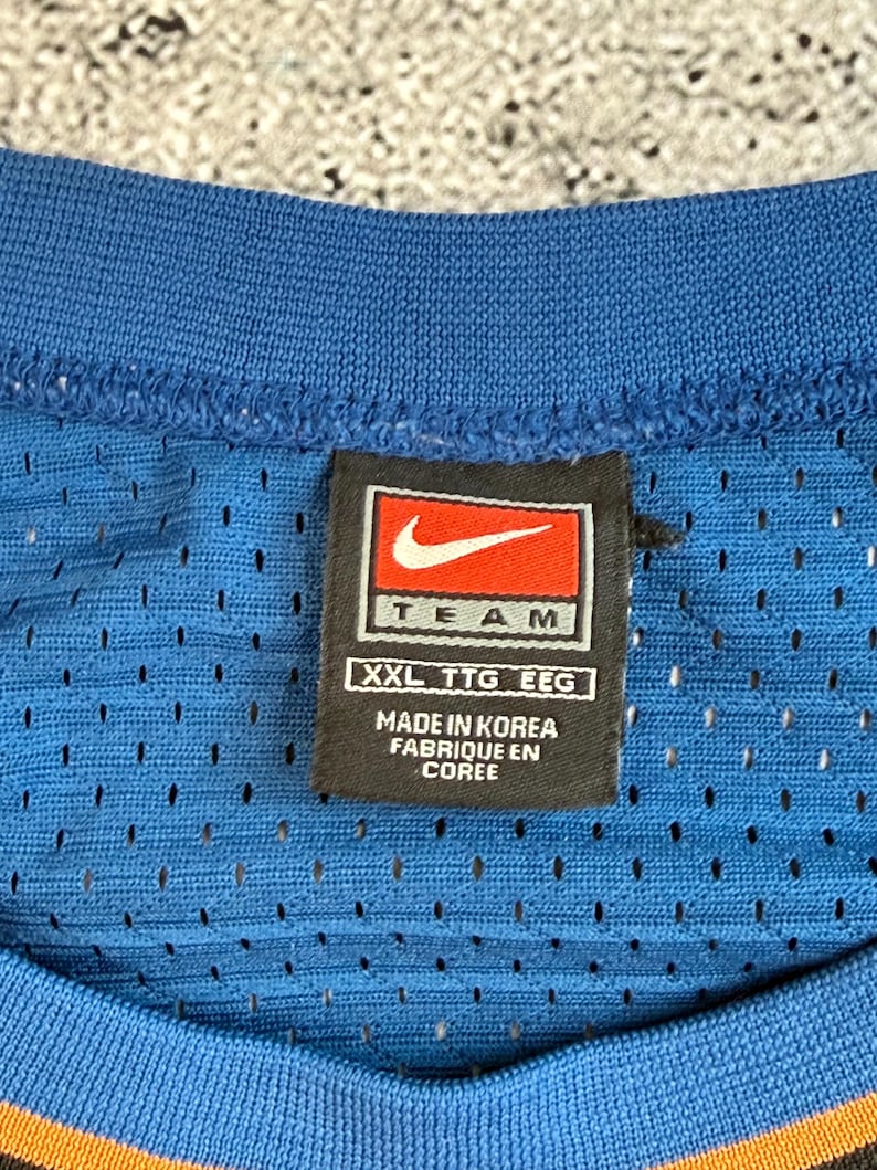 K&ouml;nnte beinhalten: Nahaufnahme eines blauen Mesh-Trikots mit einem schwarzen und roten Nike-Etikett. Auf dem Etikett steht "TEAM" mit dem Nike-Swoosh-Logo und "XXL TTG EEG". Das Trikot wird in Korea hergestellt.
