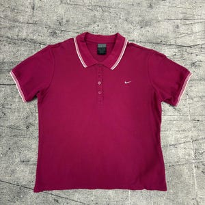 May include: A short-sleeved, magenta polo shirt with a white-trimmed collar and sleeve cuffs. The shirt features a three-button placket and a small Nike logo on the chest. The shirt is made of a soft, breathable fabric.