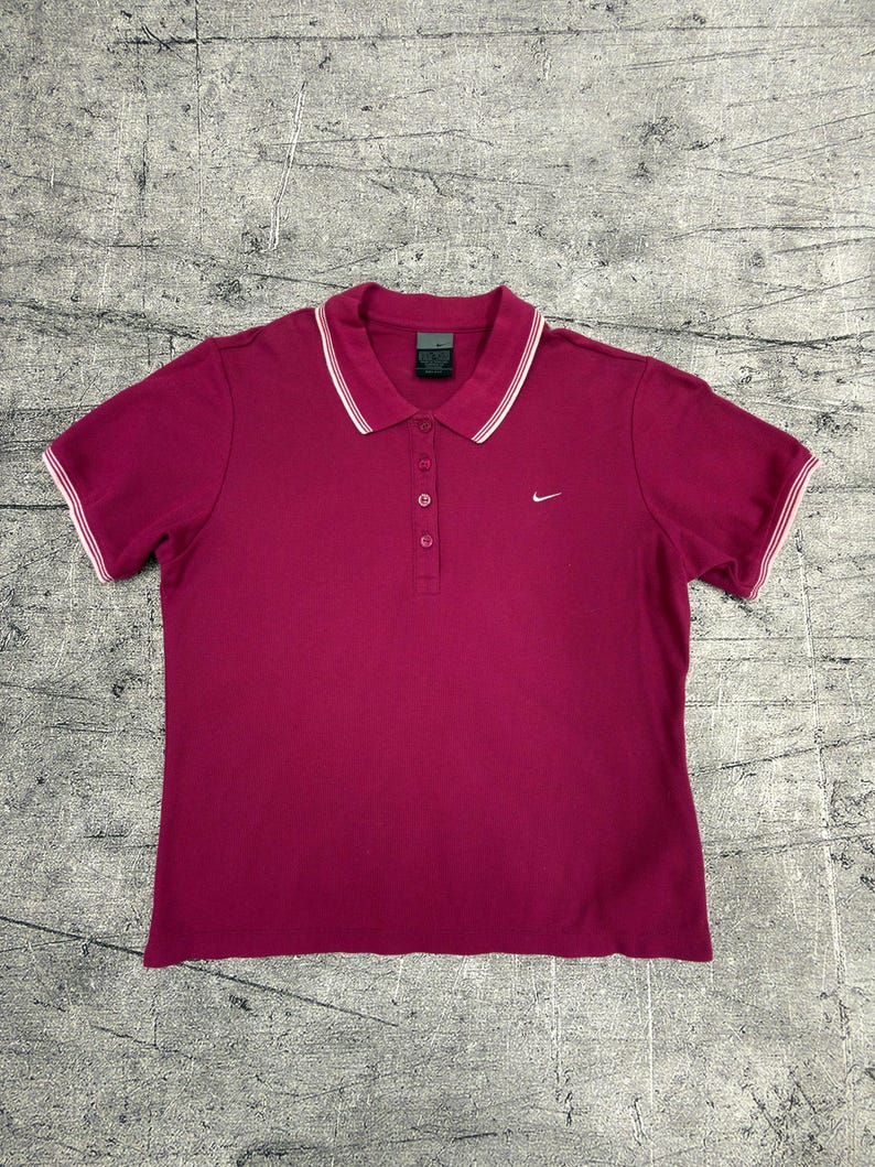 May include: A short-sleeved, magenta polo shirt with a white-trimmed collar and sleeve cuffs. The shirt features a three-button placket and a small Nike logo on the chest. The shirt is made of a soft, breathable fabric.