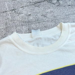 May include: White t-shirt with a crew neck and a graphic design at the bottom. The graphic features a navy blue and yellow tennis court design. The shirt is made of a soft, likely cotton material.