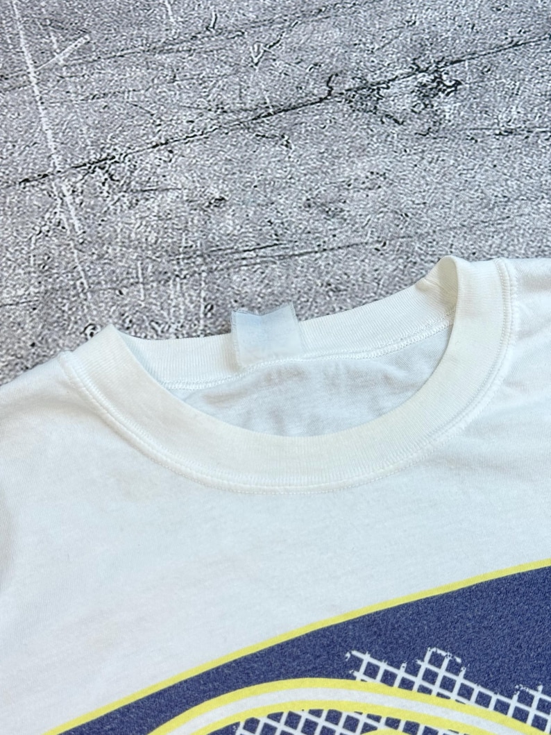 May include: White t-shirt with a crew neck and a graphic design at the bottom. The graphic features a navy blue and yellow tennis court design. The shirt is made of a soft, likely cotton material.