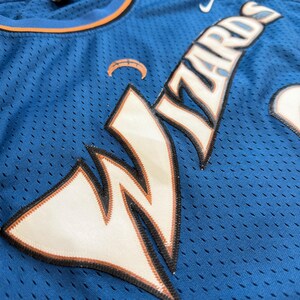 K&ouml;nnte beinhalten: Ein blaues Basketballtrikot mit dem Wort "Wizards" in wei&szlig;en und orangefarbenen Buchstaben. Das Trikot hat ein Mesh-Design und ein Nike-Logo. Das Trikot hat ein Wizards-Logo auf der Vorderseite.