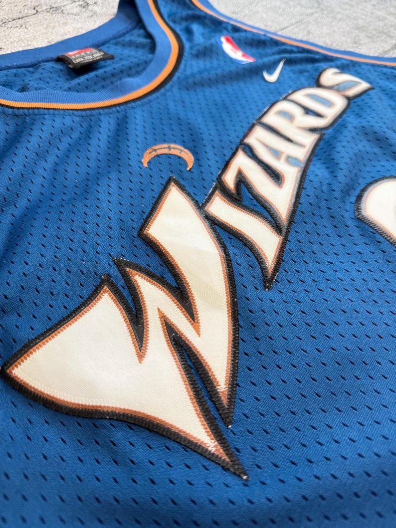 K&ouml;nnte beinhalten: Ein blaues Basketballtrikot mit dem Wort "Wizards" in wei&szlig;en und orangefarbenen Buchstaben. Das Trikot hat ein Mesh-Design und ein Nike-Logo. Das Trikot hat ein Wizards-Logo auf der Vorderseite.