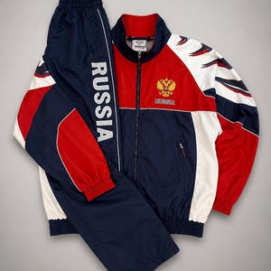 Puede incluir: Un chándal azul marino y rojo con detalles blancos. La chaqueta tiene cremallera, cuello rojo y el escudo de armas ruso. El pantalón lleva la palabra "RUSSIA" impresa verticalmente en blanco.