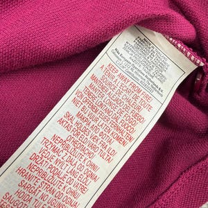 May include: Close-up of a magenta-colored knit fabric with a white tag attached. The tag has text in multiple languages, including English, French, and Spanish, with warnings about fire safety and origin of manufacture.