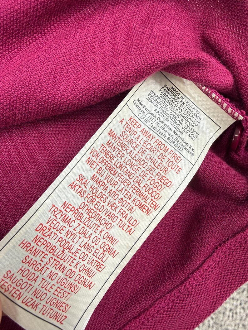 May include: Close-up of a magenta-colored knit fabric with a white tag attached. The tag has text in multiple languages, including English, French, and Spanish, with warnings about fire safety and origin of manufacture.
