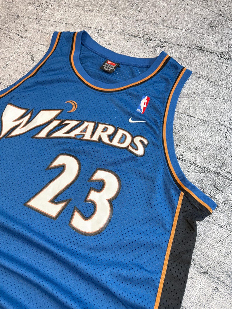 K&ouml;nnte beinhalten: Ein blaues Wizards-Basketballtrikot mit der Nummer 23 in Wei&szlig; mit kupferfarbener Umrandung. Das Trikot zeigt das Wizards-Logo und das NBA-Logo. Das Trikot hat eine Mesh-Textur und orangefarbene und schwarze Verzierungen.