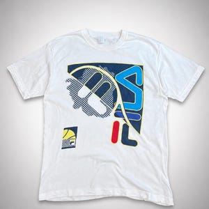 May include: White t-shirt featuring a large graphic design with the letters "FILA" in blue, red, and yellow. The design includes a tennis racket and a smaller logo in the lower left corner. The shirt has short sleeves and a crew neck.