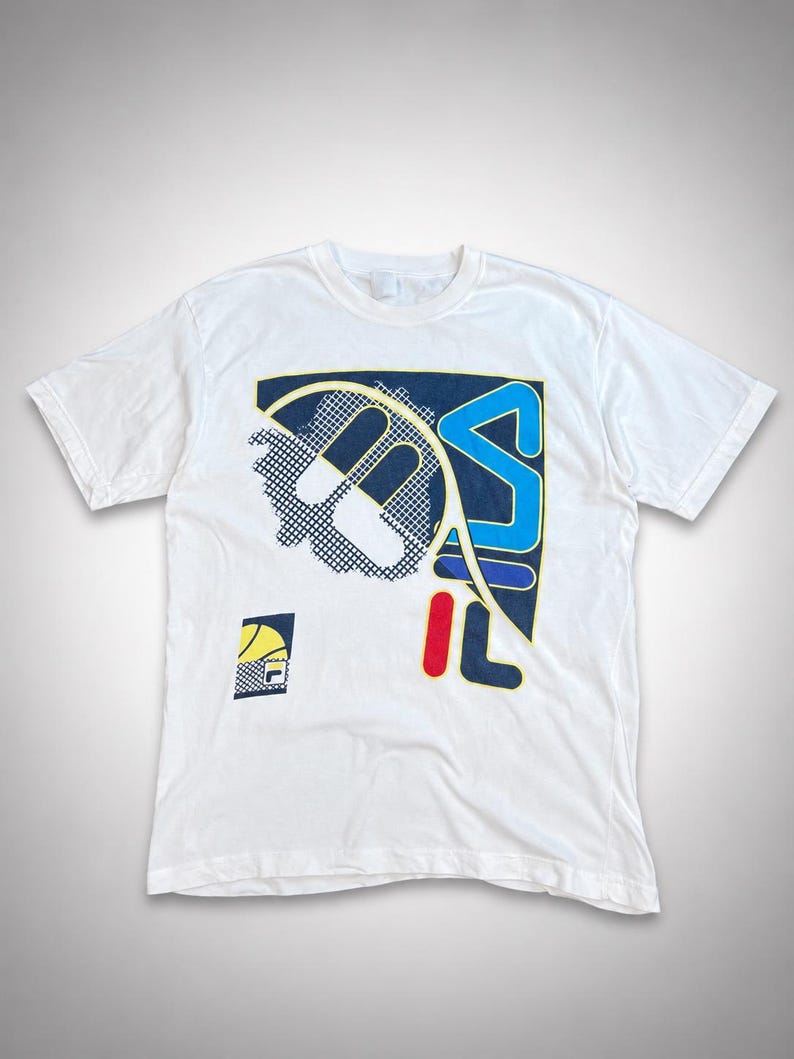 May include: White t-shirt featuring a large graphic design with the letters "FILA" in blue, red, and yellow. The design includes a tennis racket and a smaller logo in the lower left corner. The shirt has short sleeves and a crew neck.