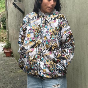 Puede incluir: Chaqueta con cremallera con un estampado repetido de cadenas doradas, joyas y las letras "SE" sobre un fondo blanco y negro. La chaqueta tiene una capucha gris y se usa con vaqueros azules.