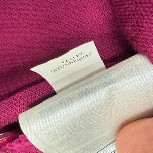 May include: Close-up of a magenta knit fabric with a white tag. The tag has the text "S40204NAY(SS) 24177A". The fabric is textured and appears to be part of a garment.
