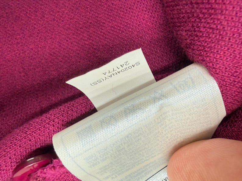May include: Close-up of a magenta knit fabric with a white tag. The tag has the text "S40204NAY(SS) 24177A". The fabric is textured and appears to be part of a garment.