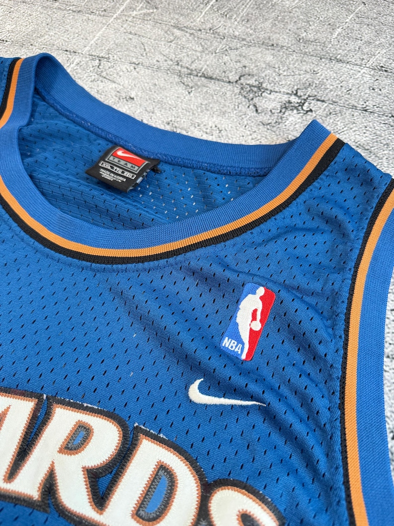 K&ouml;nnte beinhalten: Ein blaues Basketballtrikot mit orangefarbenen und schwarzen Zierleisten. Das Trikot zeigt das NBA-Logo, den Nike-Swoosh und das Wort "WIZARDS" in Wei&szlig;. Das Trikot hat einen V-Ausschnitt und besteht aus atmungsaktivem Material.