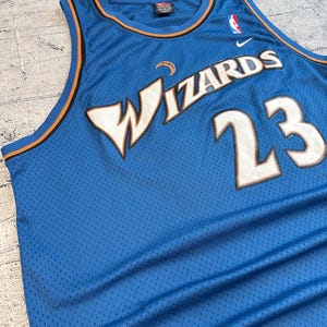 K&ouml;nnte beinhalten: Ein blaues Basketballtrikot aus Mesh mit dem Wort "WIZARDS" in Wei&szlig; und der Nummer "23" in Wei&szlig; mit goldener Umrandung. Das Trikot hat orangefarbene und blaue Verzierungen und ein Nike-Logo.
