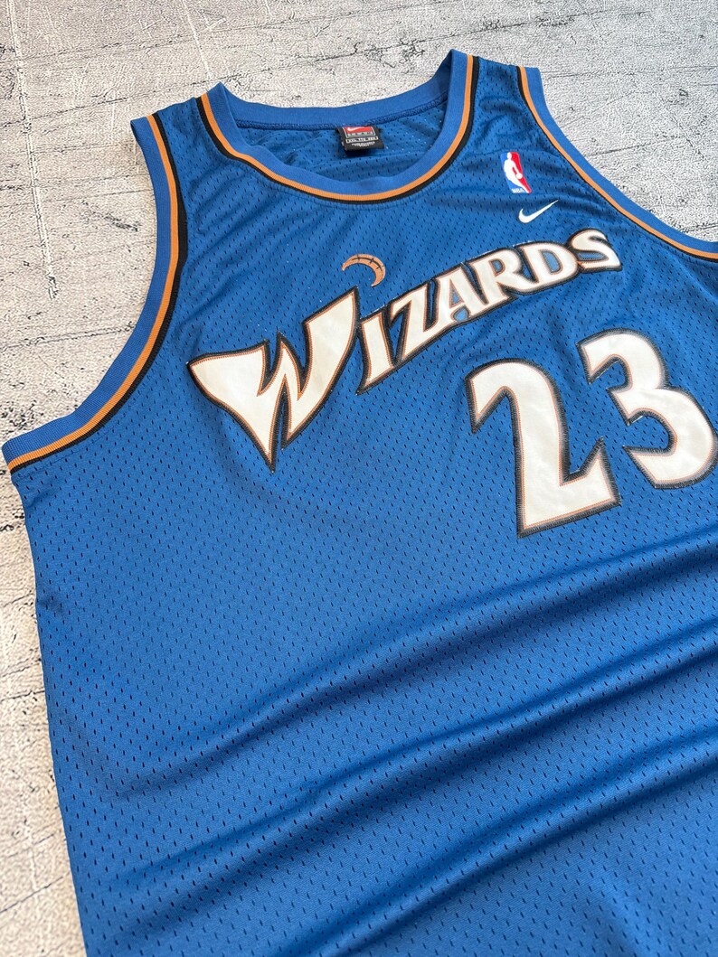 K&ouml;nnte beinhalten: Ein blaues Basketballtrikot aus Mesh mit dem Wort "WIZARDS" in Wei&szlig; und der Nummer "23" in Wei&szlig; mit goldener Umrandung. Das Trikot hat orangefarbene und blaue Verzierungen und ein Nike-Logo.