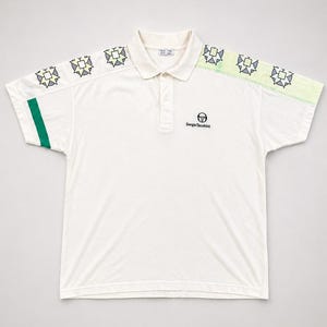 Puede incluir: Polo de color crema con cuello y mangas cortas. La camisa presenta una franja verde en la manga izquierda y un estampado geométrico en los hombros. El logotipo de Sergio Tacchini está bordado en el pecho.
