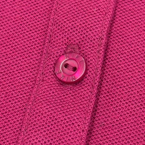 May include: Close-up of a magenta-colored knit fabric with three matching buttons. The buttons are round and have a subtle sheen, with the brand name subtly embossed on each. The fabric has a textured appearance.