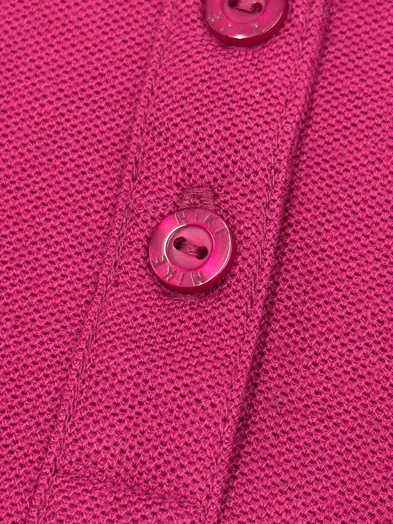 May include: Close-up of a magenta-colored knit fabric with three matching buttons. The buttons are round and have a subtle sheen, with the brand name subtly embossed on each. The fabric has a textured appearance.