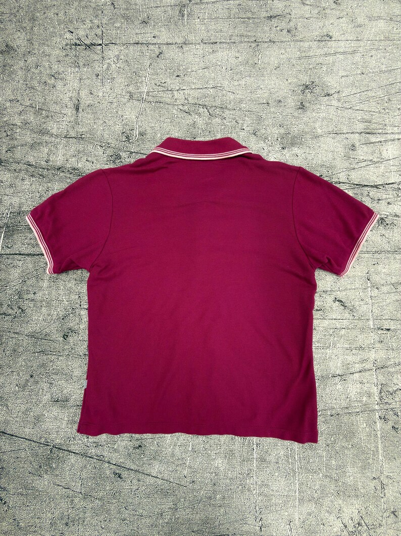 May include: A short-sleeved polo shirt in a deep magenta color. The shirt features a white collar and white trim along the sleeves. The back view of the shirt is shown, highlighting the classic polo design.