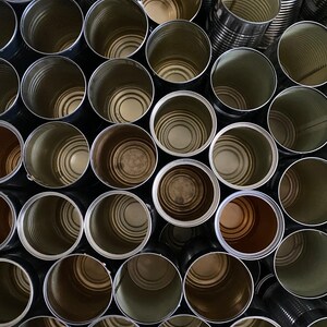 May include: Overhead view of a collection of empty metal cans. The cans are cylindrical with a silver exterior and a visible interior. Some cans have a darker, brownish hue inside. The arrangement is dense, with the open tops of the cans facing upwards.