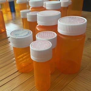 May include: A collection of empty orange plastic prescription pill bottles with white child-resistant caps. The caps feature printed instructions for opening and closing the containers. The bottles vary in size and are arranged in a group.