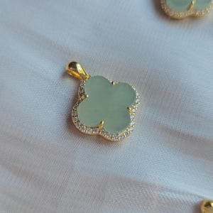 May include: A gold-coloured clover pendant with a pale green centre stone and a border of small, clear crystals. The pendant has a loop at the top for a chain. The background is a light, neutral fabric.