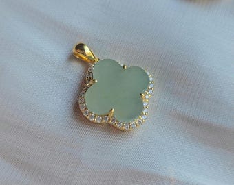 Puede incluir: Un colgante de trébol dorado con una piedra central verde claro y un borde de pequeños cristales transparentes. El colgante tiene un lazo en la parte superior para una cadena. El fondo es una tela clara y neutra.