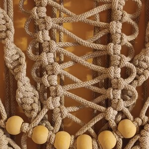 May include: A beige macrame wall hanging with wooden beads. The macrame is made with thick rope and has a geometric pattern. The wooden beads are round and have a natural finish.