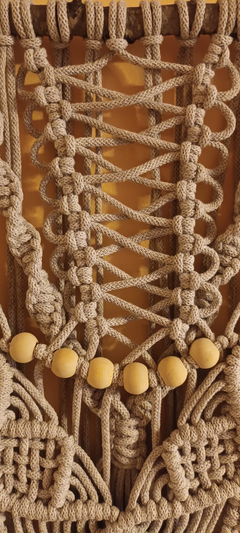 May include: A beige macrame wall hanging with wooden beads. The macrame is made with thick rope and has a geometric pattern. The wooden beads are round and have a natural finish.