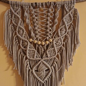 May include: A beige macrame wall hanging with wooden beads and fringe. The hanging is attached to a branch.
