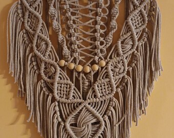 Op de afbeelding: Een beige macramé wandhanger met houten kralen en franjes. De hanger is bevestigd aan een tak.