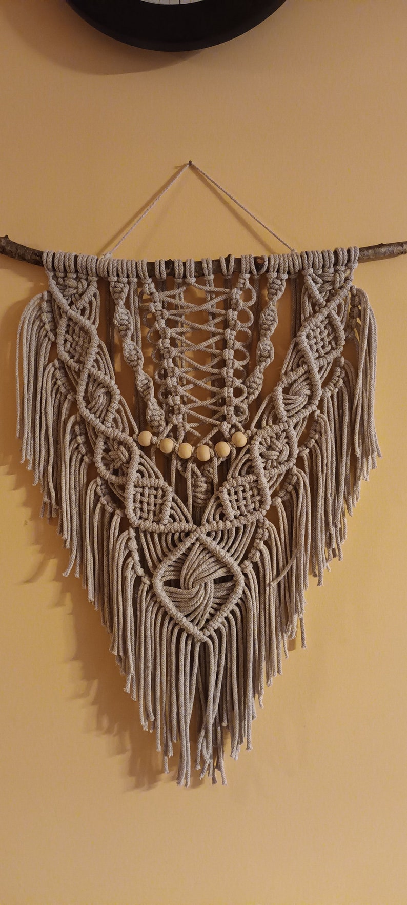 May include: A beige macrame wall hanging with wooden beads and fringe. The hanging is attached to a branch.