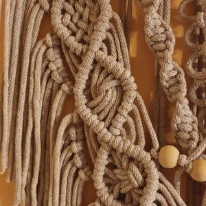 May include: A beige macrame wall hanging with wooden beads. The macrame is made with a variety of knots and patterns, creating a unique and intricate design.