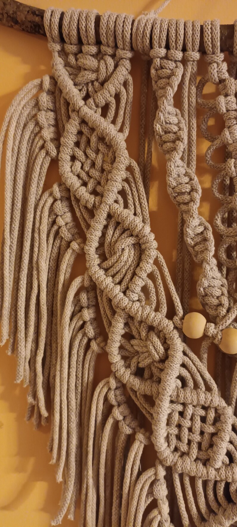 May include: A beige macrame wall hanging with wooden beads. The macrame is made with a variety of knots and patterns, creating a unique and intricate design.
