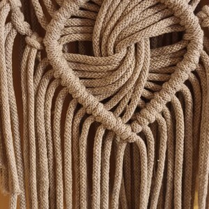 May include: A beige macrame wall hanging with a woven design and long fringe. The fringe is made of the same beige cord as the wall hanging.
