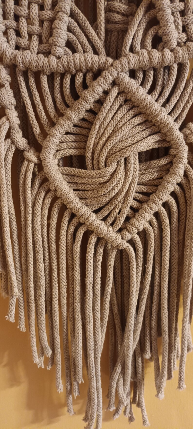 May include: A beige macrame wall hanging with a woven design and long fringe. The fringe is made of the same beige cord as the wall hanging.